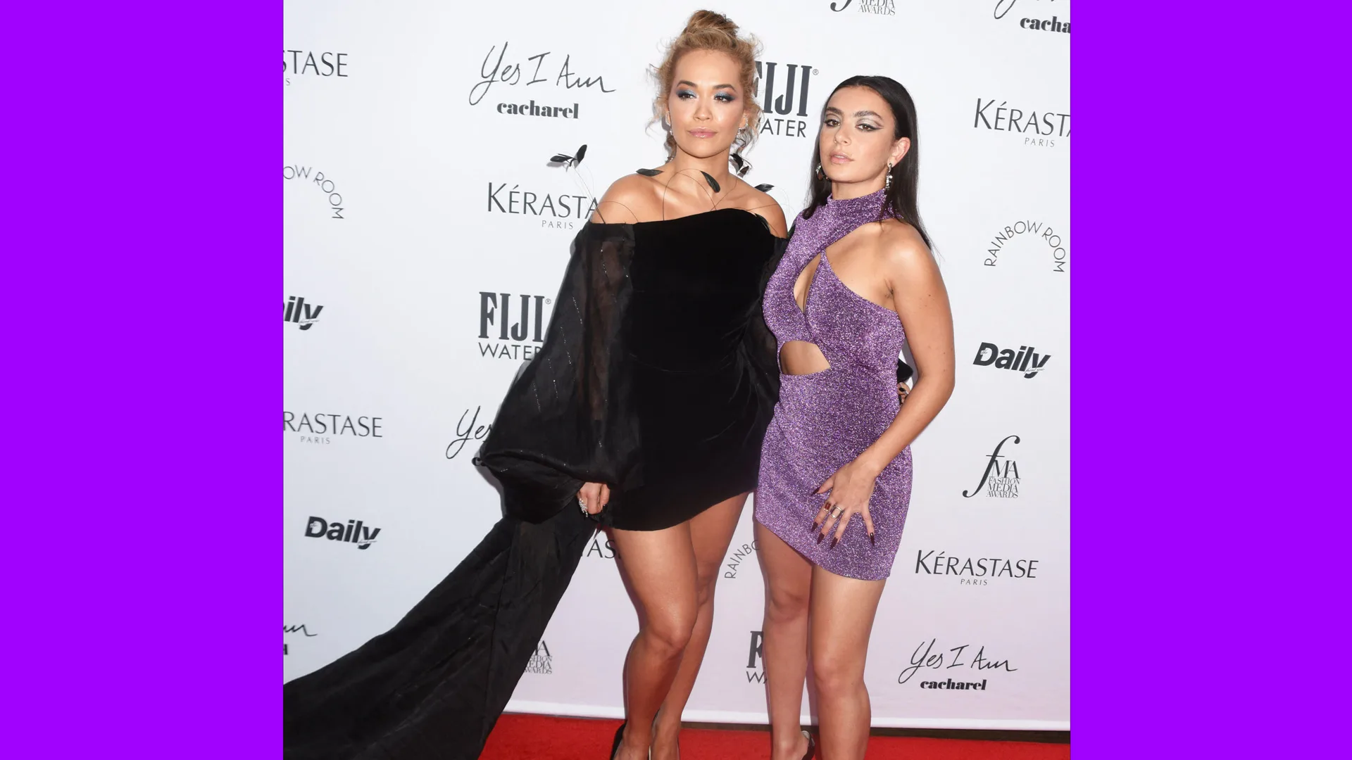 A photo of Charli XCX and Rita Ora on the red carpet. Rite is wearing a black dress with an oversized sleeve that has a train trailing on the carpet. Charli is wearing a purple glitter dress. They are stood against a white wall with brand names.